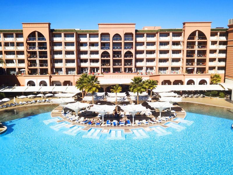 Groot hotel met centraal zwembad, parasols en ligstoelen onder een heldere hemel.