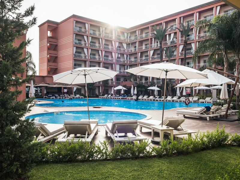 Groot hotelzwembad met ligbedden en parasols, omgeven door een gebouw met meerdere verdiepingen en palmbomen.
