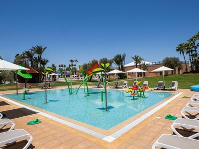 Ein großer Außenpool mit Wasserspielen, umgeben von Liegestühlen und Sonnenschirmen unter blauem Himmel.