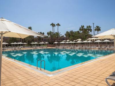 Großer, rechteckiger Pool mit blauen Wasserspiegel, umgeben von beigen Sonnenschirmen und Liegestühlen.