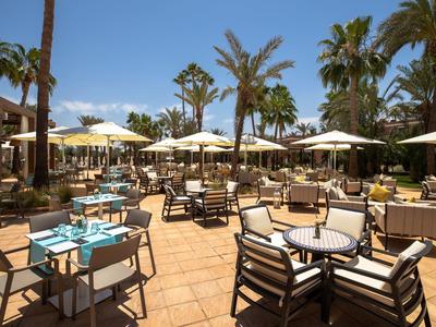 Freiluftrestaurant mit Tischen, Stühlen, Sonnenschirmen und Palmen bei sonnigem Wetter.