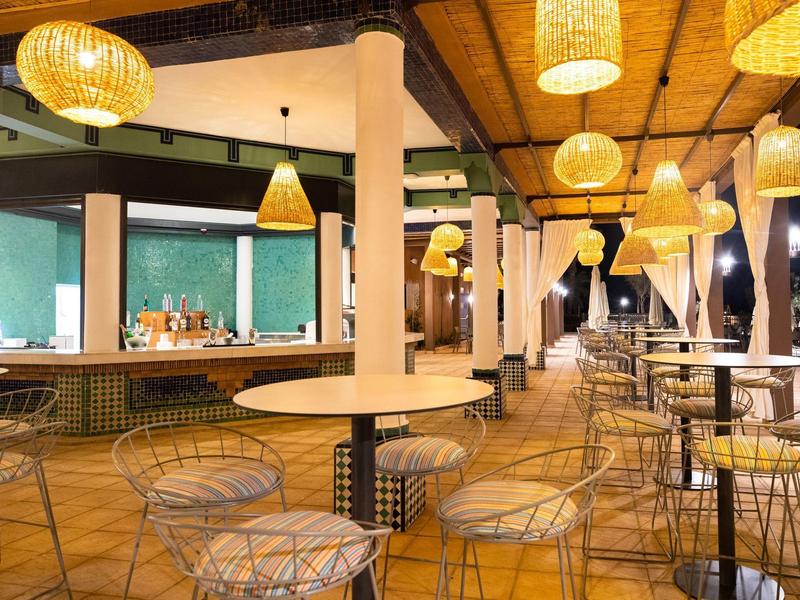 Freiluftbar mit hohen Tischen, Metallstühlen und warmen, gelben Hängeleuchten unter Holzdach.