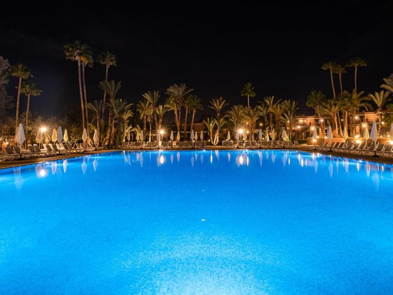 Großer Pool mit blau beleuchtetem Wasser, umgeben von Palmen und beleuchteten Sonnenschirmen bei Nacht.