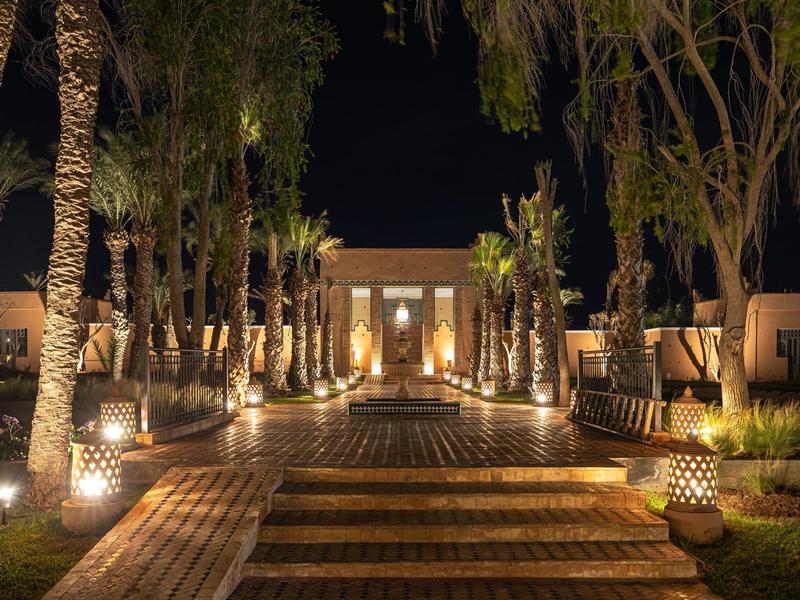 Beleuchteter Weg mit Palmen und Laternen führt zu einem Gebäude in warmem Licht bei Nacht.