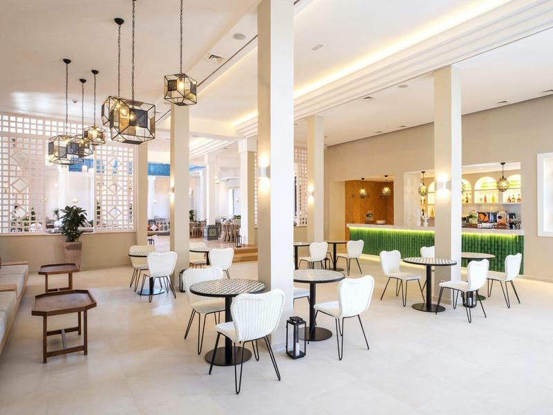 Moderner heller Gastraum mit weißen Stühlen, Holz- und Glaselementen, Pflanzen und Deckenbeleuchtung.