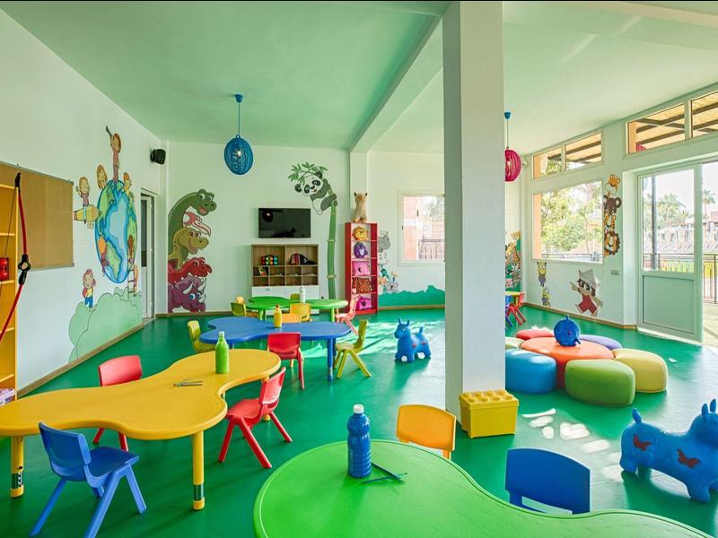 Bunter Kindergartenraum mit kleinen Tischen, Stühlen und Spielzeugfiguren auf hellem Boden.