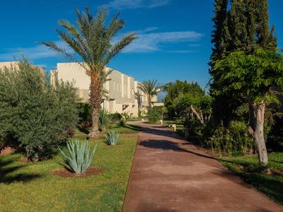 Un sentiero lastricato attraversa un giardino verde con palme e cespugli verso edifici sotto un cielo azzurro.