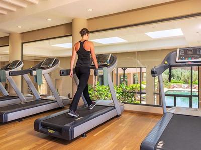 Woman walks on treadmill in gym with large windows overlooking a garden.