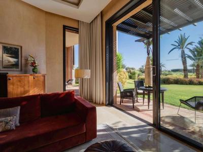 Bright hotel lounge with red sofa, wooden furniture, and large glass sliding door to garden with palms.
