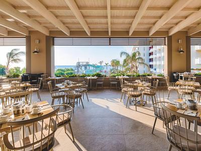 Helles, modernes Restaurant mit Holztischen und Stühlen mit Blick auf Palmen und Meer.