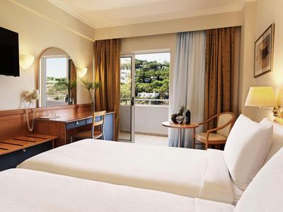 Helles Hotelzimmer mit zwei Betten, Schreibtisch, Stuhl und Balkon mit Blick ins Grüne.