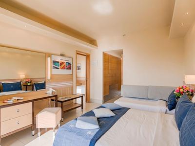 Hell eingerichtetes Hotelzimmer mit Bett, Schreibtisch, Hocker und Sofa.