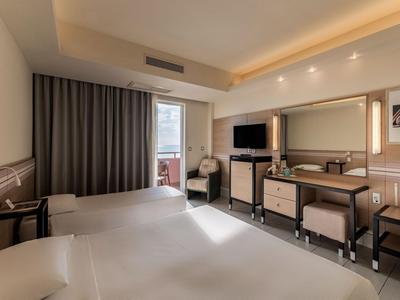 Helles Hotelzimmer mit zwei Betten, Schreibtisch, Fernseher und Balkon mit Blick nach draußen.
