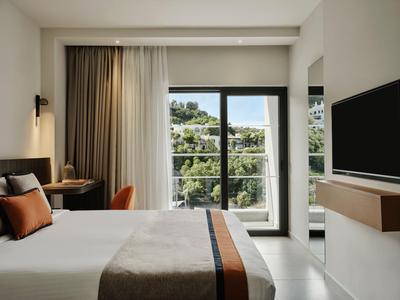 Modernes Hotelzimmer mit Doppelbett, Schreibtisch, Fernseher und Balkon mit Blick auf Bäume.