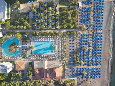 Luftaufnahme eines Resorts mit Pools, Sonnenschirmen und Liegen am Strand und Palmen.