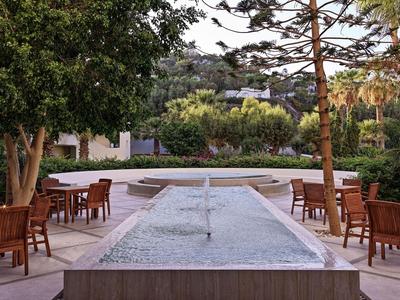 Springbrunnen mit Wasserstrahl umgeben von Holztischen und Stühlen in einem Garten.