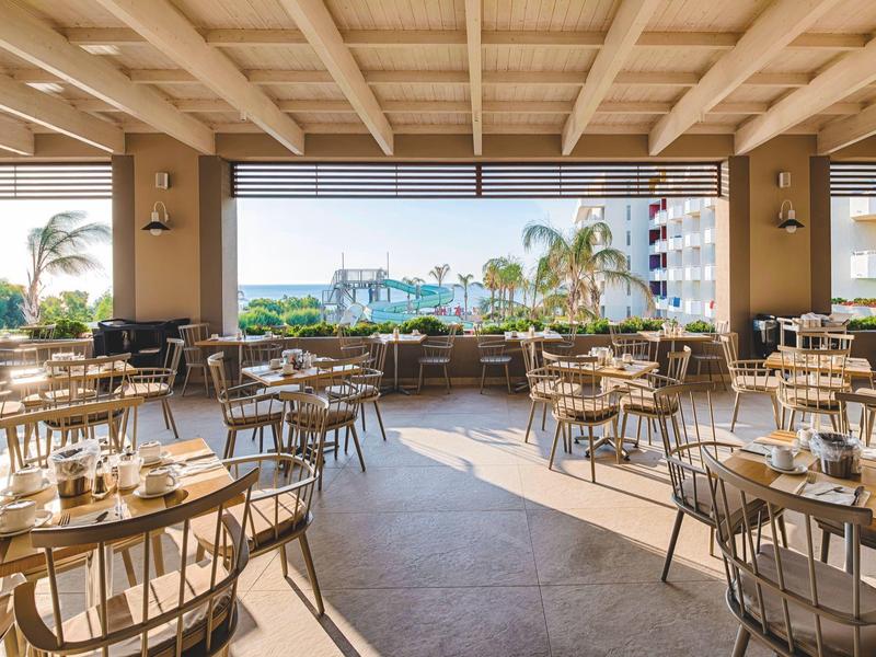 Helles, modernes Restaurant mit Holzdecken, runden Tischen und Blick auf Palmen und Meer.