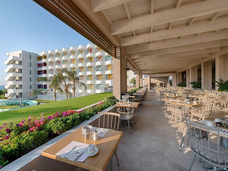 Moderner Hotelbalkon mit vielen Tischen und Stühlen, Blick auf weißen Gebäudekomplex und Garten.