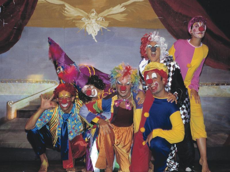 Bunte Clowns posieren auf Bühne mit rotem Vorhang und leuchtendem Vogel an der Decke.