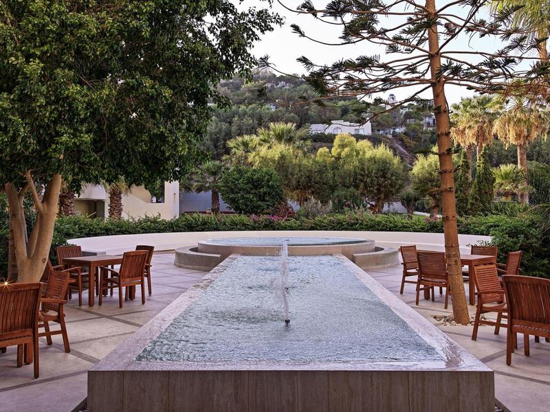 Ein ruhiger Außenbereich mit Springbrunnen, umgeben von Tischen und Stühlen in einem bewaldeten Hotelgarten.