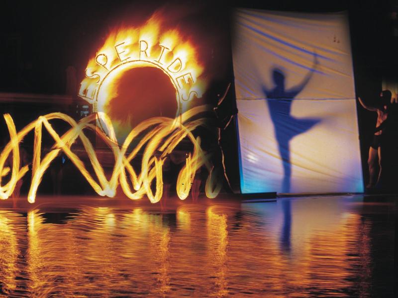 Bühnenbild mit Schatten einer Tänzerin vor weißem Vorhang und buntem Lichtspiel am Wasser.