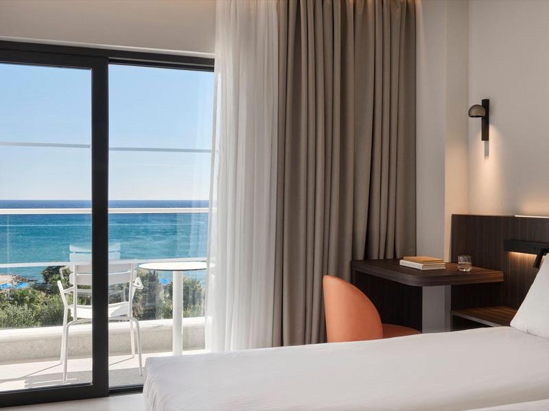 Hotelzimmer mit Bett, Schreibtisch und Meerblick durch bodentiefe Fenster.