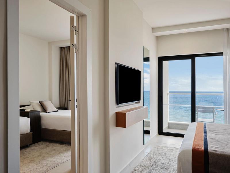 Helles Hotelzimmer mit Bett, Fernseher an der Wand und Balkon mit Meerblick.
