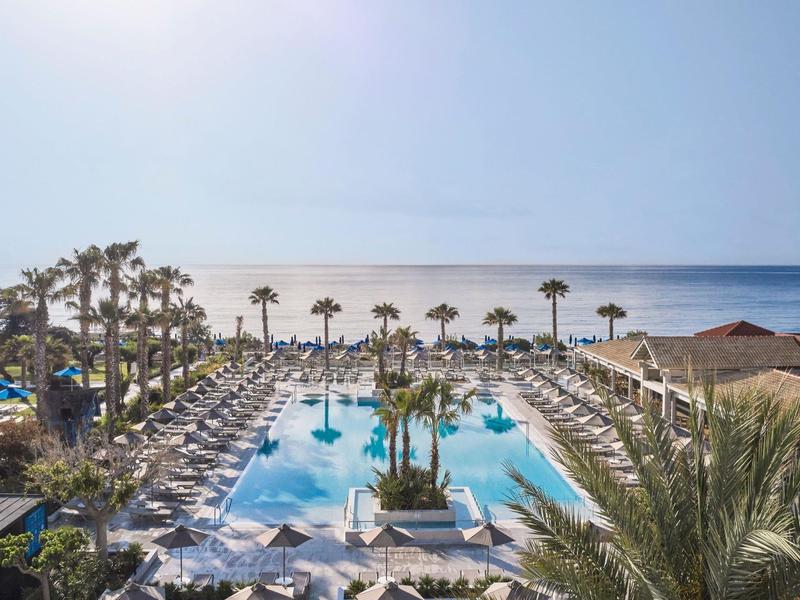 Blick auf einen Pool mit Palmen und Sonnenliegen am Meer an einem sonnigen Tag.