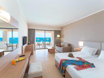 Helles Hotelzimmer mit Doppelbett, Sofa, Schreibtisch, Balkon und Meerblick bei Tageslicht.