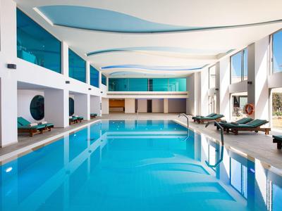 Großes Hallenbad mit blauem Wasser, weißen Wänden, Liegestühlen und großen Fenstern.