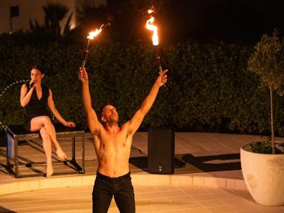 Ein shirtloser Mann führt eine Feuershow draußen auf, während eine Frau auf einer Bank zuschaut.