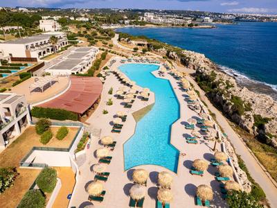Großer Pool mit Sonnenschirmen und Liegen neben Küste und Felsen, Hotel und blauer Himmel.
