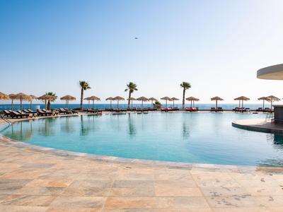 Runder Pool mit Liegestühlen und Sonnenschirmen am Meer unter blauem Himmel