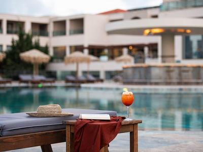 Poolbereich mit Liegestuhl, Strohhut, Buch, rotem Tuch und Cocktailglas im Vordergrund.