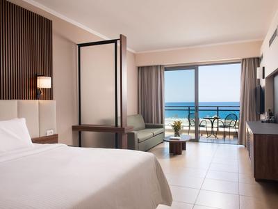 Helles Hotelzimmer mit Bett, Sofa, Balkon und Meerblick bei Tageslicht.