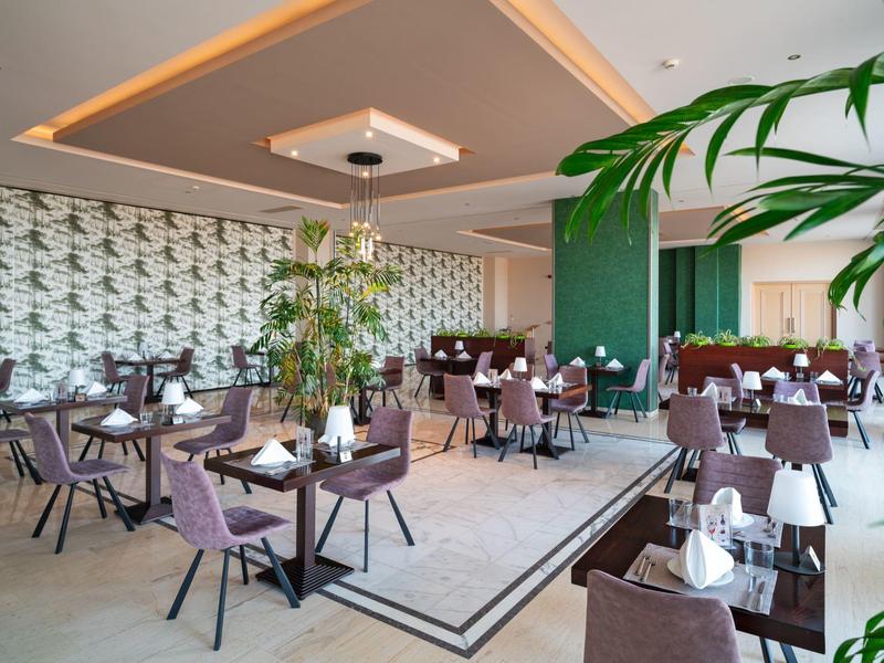 Modernes Restaurant mit lila Stühlen, gedeckten Tischen und grünen Wänden, umgeben von Pflanzen.