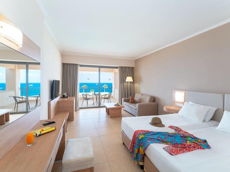 Helles Hotelzimmer mit Doppelbett, Sofa, Schreibtisch, Balkon und Meerblick bei Tageslicht.