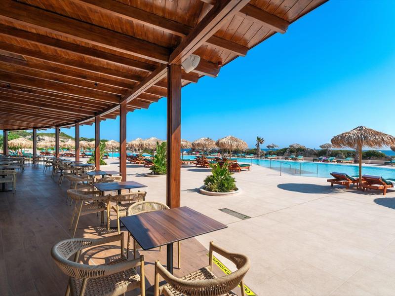 Große Terrasse mit Holztischen und Stühlen neben einem Pool mit Sonnenschirmen und blauem Himmel.