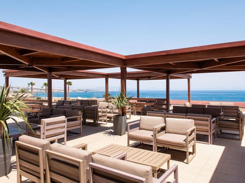 Dachterrasse mit Holzüberdachung, bequemen Sitzgruppen und Blick aufs Meer unter klarem Himmel.