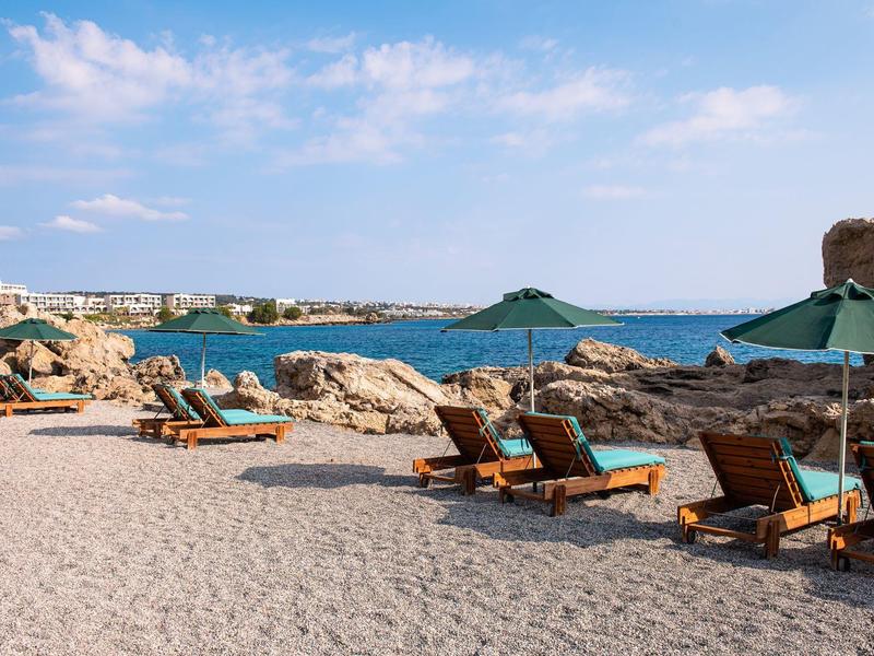 Strand mit Liegestühlen, grünen Sonnenschirmen und felsiger Küste unter blauem Himmel.
