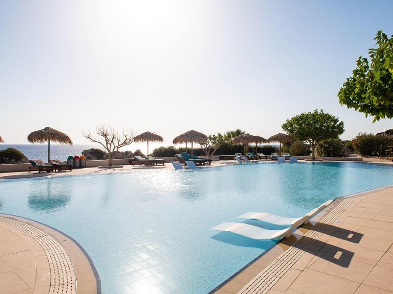 Großer Pool mit klarem Wasser, Sonnenschirmen und Bäumen unter hellem Himmel am Nachmittag.