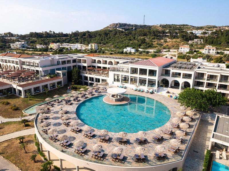 Großes Hotel mit rundem Außenpool, vielen Sonnenliegen und umliegenden Gebäuden in Hügelumgebung.