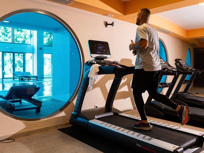 Mann joggt auf Laufband in modernem Fitnessraum mit großem Rundfenster und natürlichem Licht.