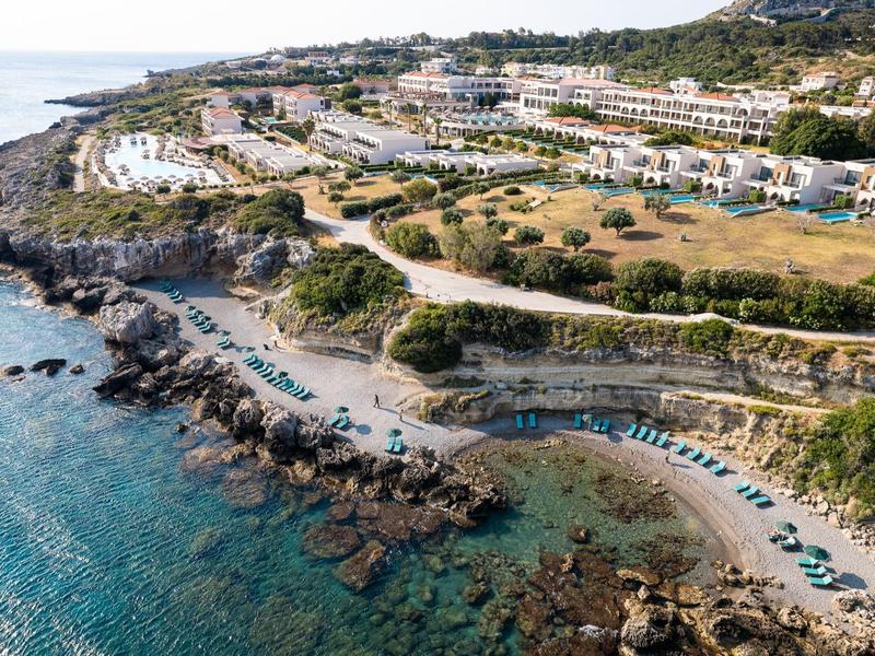 Küstenlandschaft mit klaren blauen Wasser, Felsenstrand und weißen Gebäuden auf hügeligem Gelände.