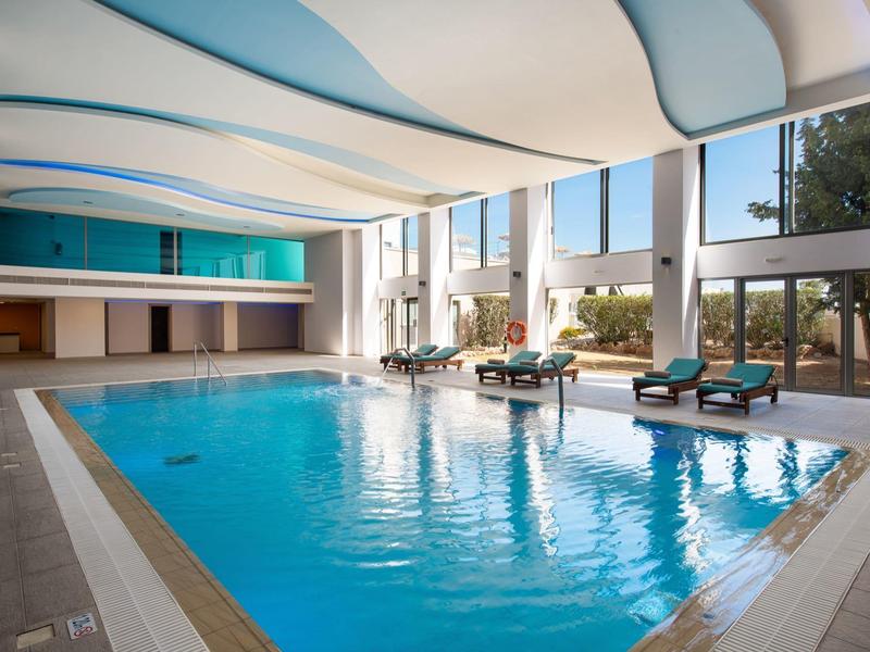 Hallenbad mit hellblauem Wasser, Liegestühlen am Rand und großen Fenstern für Tageslicht.