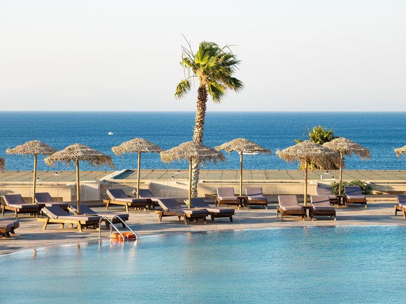 Ein Pool mit Liegen und Sonnenschirmen aus Stroh am Meer, eine Palme im Zentrum.