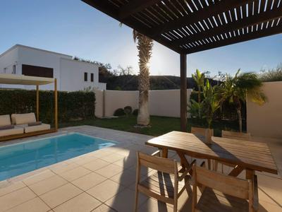 Zona de piscina con mesa de madera y sillas bajo una pérgola, rodeada de jardín y muro.