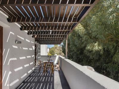 Balcón con techo de madera, patrón de sombra en el suelo, jardín con árboles junto a muro blanco