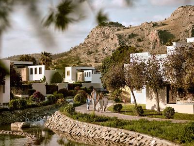 Paesaggio con edifici bianchi e montagne sullo sfondo, escursionisti lungo un sentiero accanto a un ruscello.