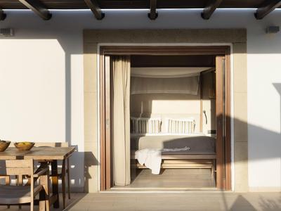 Camera aperta con porta in legno e tende, a sinistra tavolo in legno con sedie sulla terrazza.